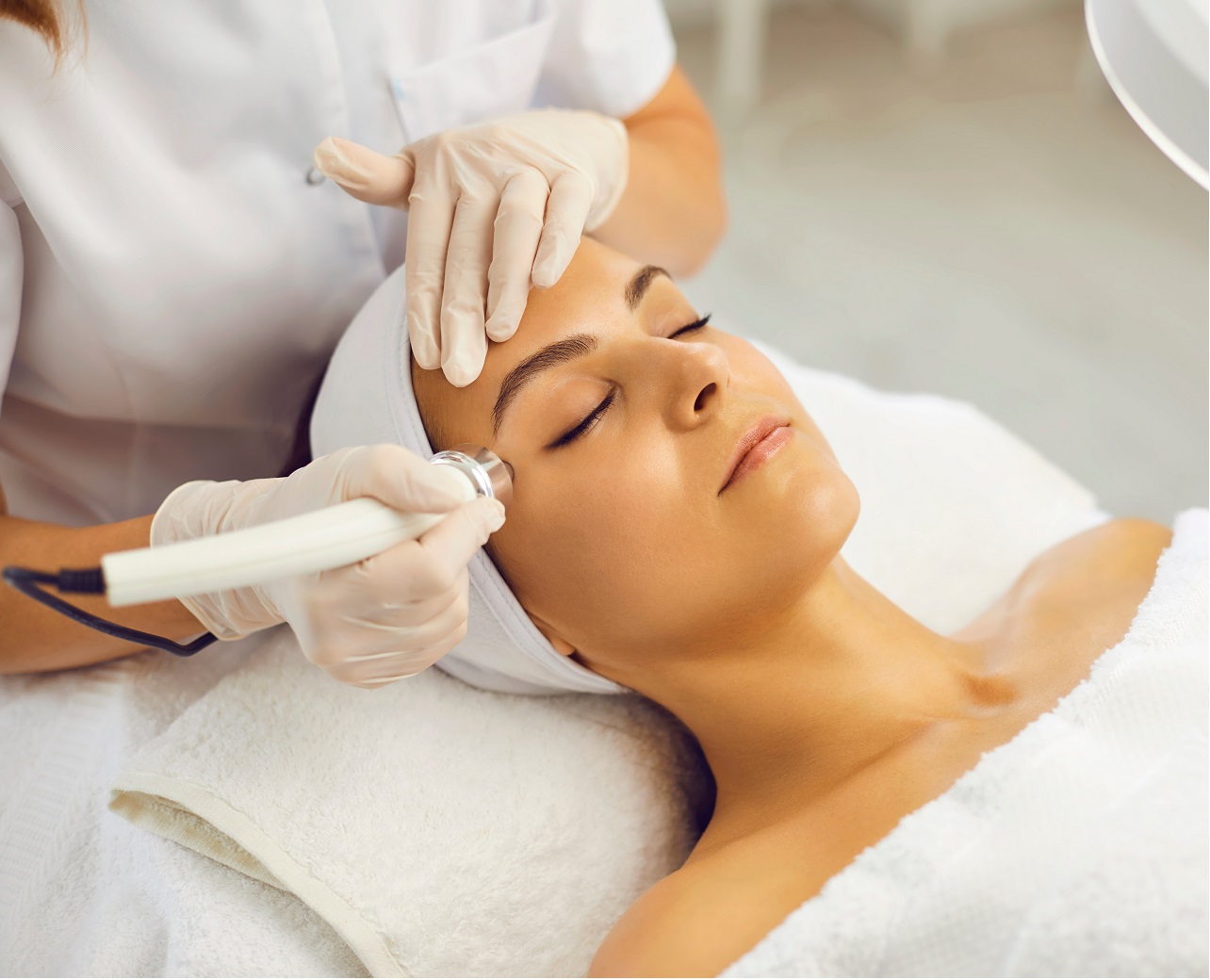 Relaxed young woman with eyes closed getting procedure of vacuum facial cleaning from cosmetologist in gloves in beauty spa salon. Facial treatment, skincare, cosmetology concept | Aesthetic Center of Richmond Dermatology in Medical spa in Glen Allen, VA
